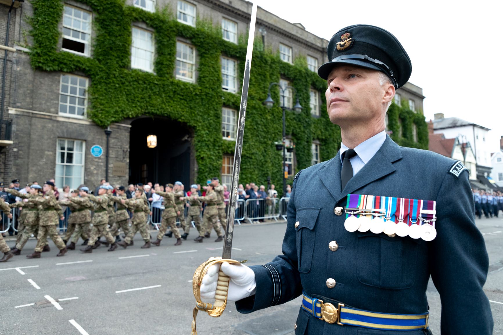 RAF Aviator holds Pooley Sword with parade in background, through the streets.